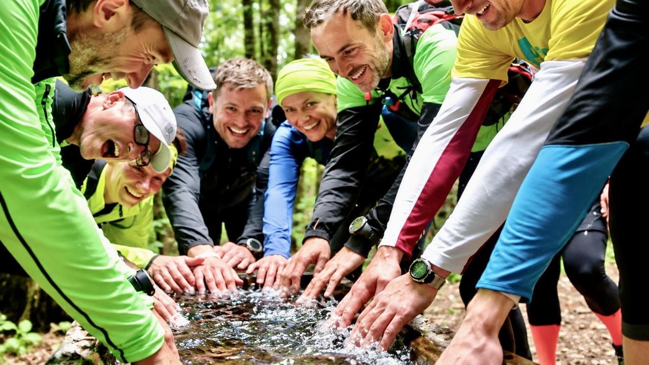 Pavel Hruška mit einem Laufteam im Wald, alle lachen und tauchen die Hände in eine Quelle